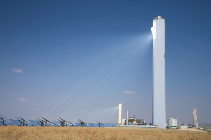 Sun rays visible in hazy atmosphere reflected from an array of heliostats toward a central tower solar power plant. (Source: © Q / stock.adobe.com)